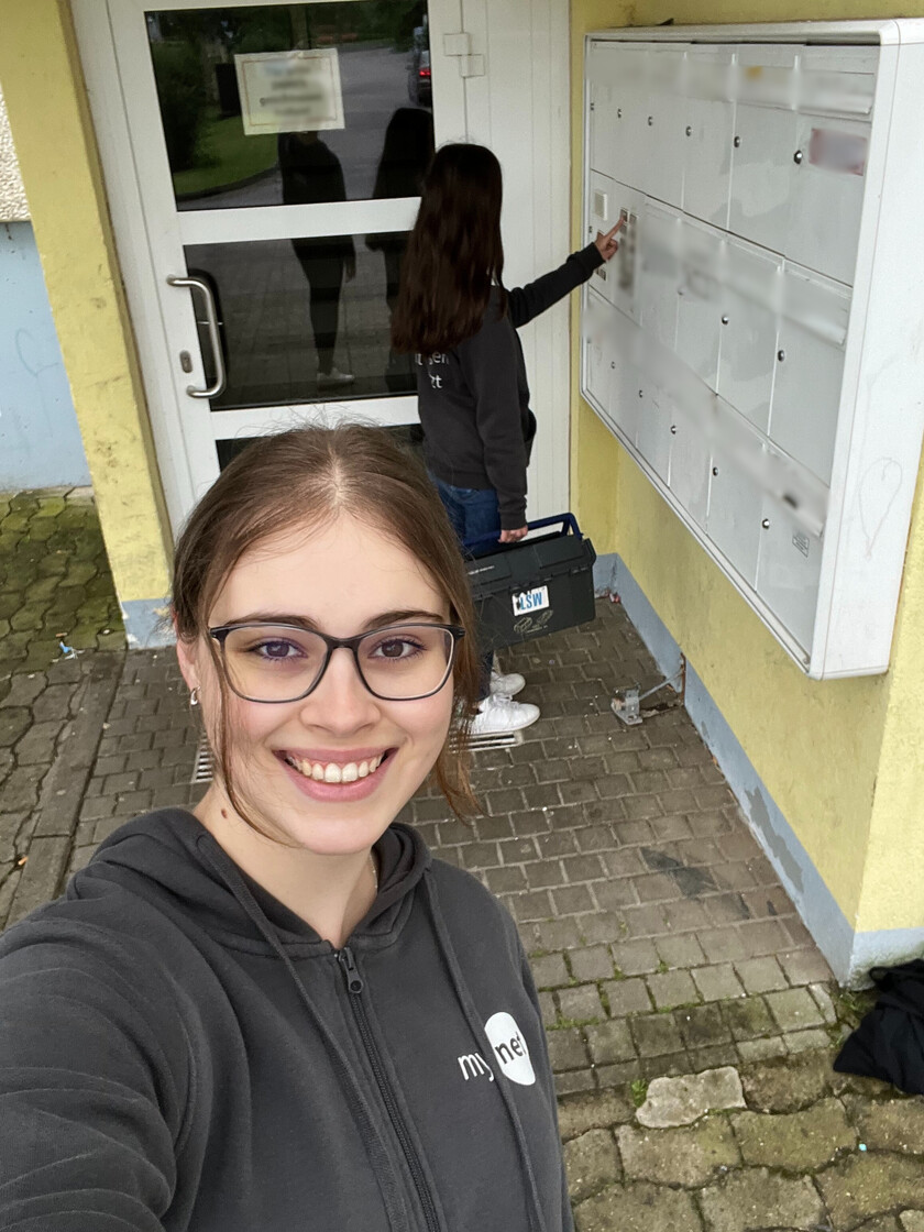 Vorne steht ein dunkelhaariges Mädchen mit Brille und lächelt in die Kamera. Hinten steht ein zweites Mädchen mit denkelen, offenen Harren und klingelt. Beide stehe vor einem Hauseingang eines Mehrfamilienhauses.