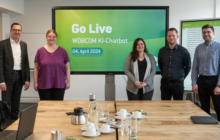 Das Projekt Team steht vor einem großen Bildschirm mit der Aufschrift "Go Live WOBCOM KI-Chatbot 04. April 2024" und freuen sich über die Liveschaltung des KI-Chatbots. Vor dem Bildschirm stehen v.l. Dalibor Dreznjak, Jessica Klapproth, Chantal Heinemann, Tobias Malonnek und Nikola Simovic.