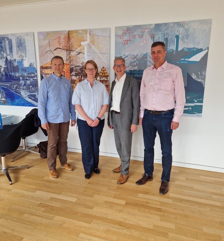 Kai Kronschnabel (1. stellvertretender Vorsitzender des Aufsichtsrats) steht zusammen mit Birte Fleischer (Vorständin Stadtwerke Wolfsburg AG), Markus Alisch (Aufsichtsratsvorsitzender) und Kai Ritter (2. stellvertretender Vorsitzender des Aufsichtsrats) zusammen vor einer weißen Wand mit bunten Bildern. Alle schauen und lächeln in die Kamera.