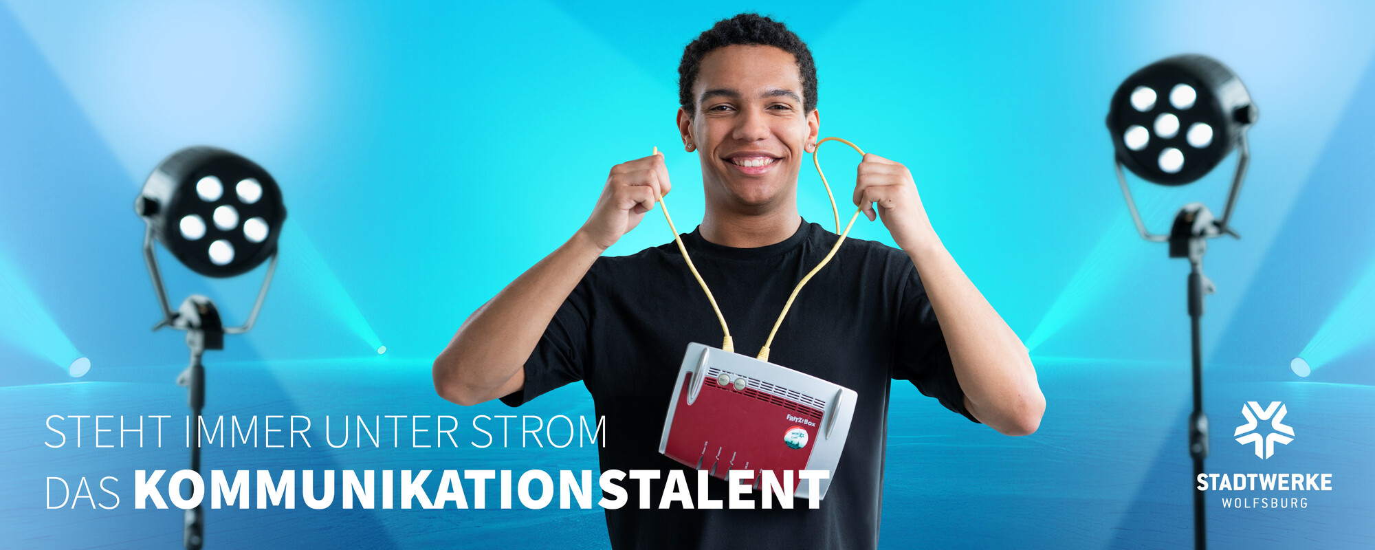 Ein junger Mann hält einen Router an zwei Netzwerkkabeln vor seinen Körper. Text: Steht immer unter Strom - Das Kommunikationstalent