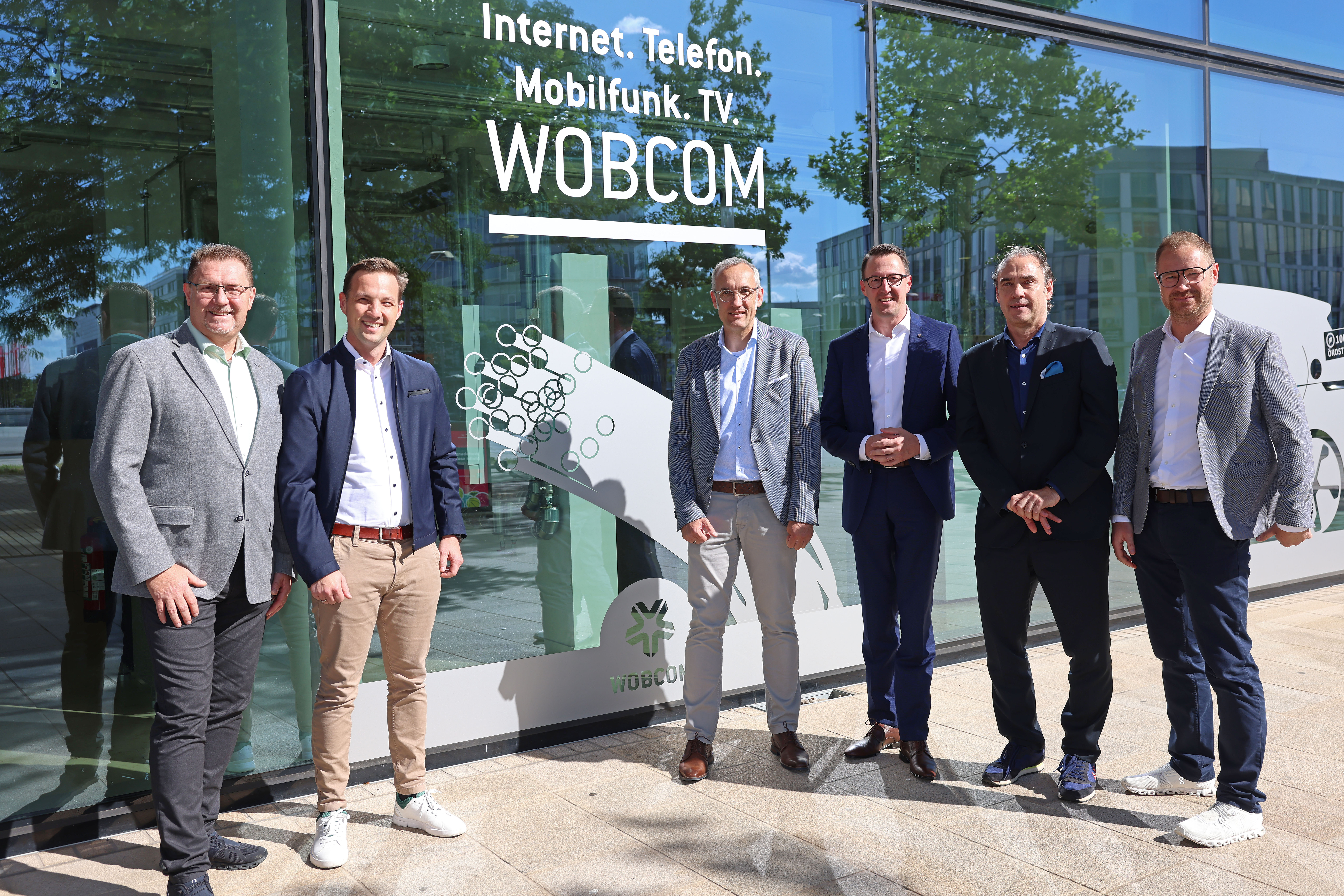 v.l. Thomas Schulz (WOBCOM Abteilungsleiter Vertrieb und Kundenservice), Benedikt Brandau (Deutsche Glasfaser Bereichsleiter Wholesale), Kai Scholz (Deutsche Glasfaser Business Director Wholesale), Dalibor Dreznjak (WOBCOM Geschäftsführer) Frank Pikelj (Deutsche Glasfaser Abteilungsleiter Key Account Management Wholesale) und Anatoli Seliwanow (WOBCOM Abteilungsleiter Betrieb) stehen vor einem großen Fenster mit der Beklebung "Internet. Telefon. Mobilfunk. TV. Wobcom und einem Glasfaser Symbol. Alle lächeln in die Kamera.
