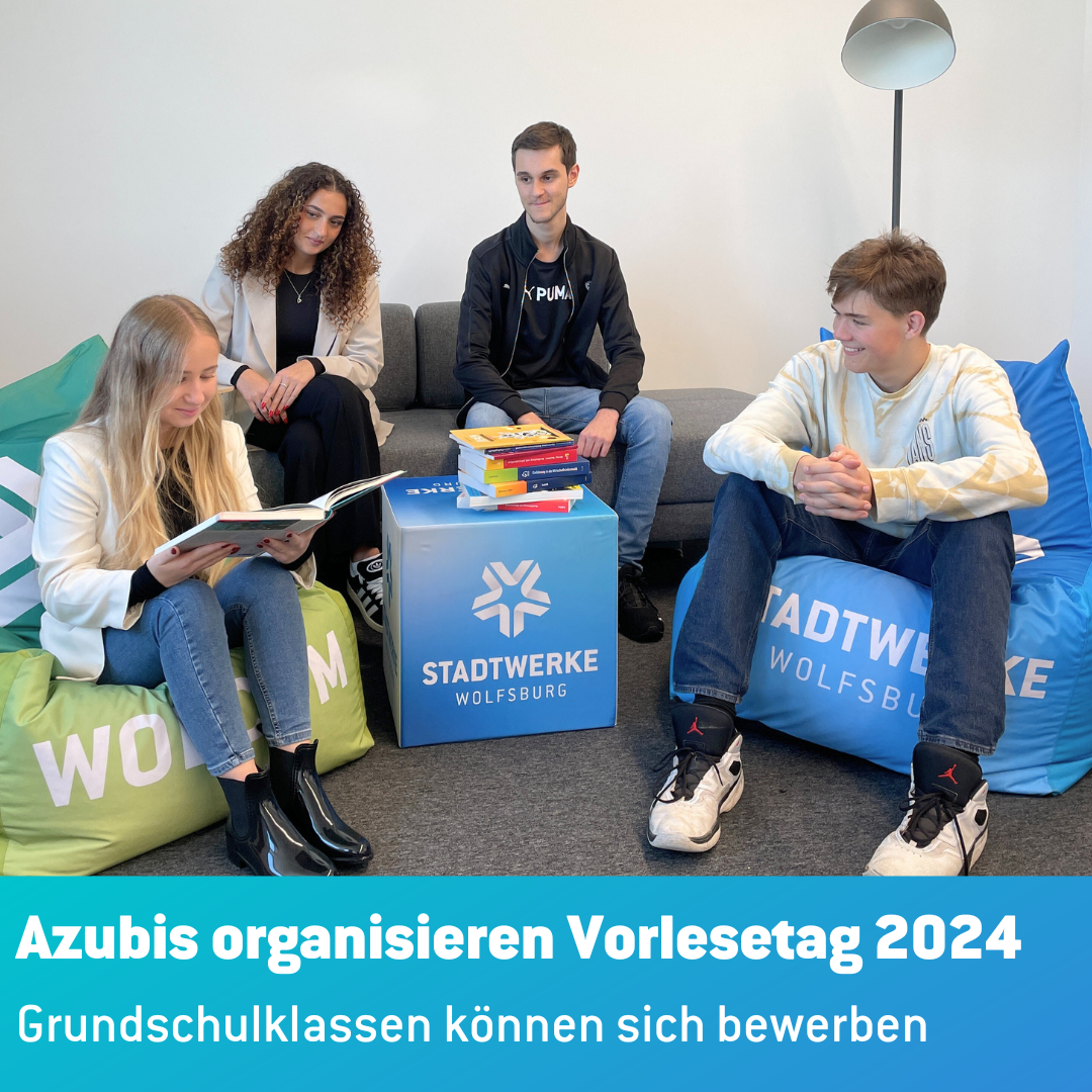 Vier Azubis sitzen zusammen. Zwei auf einer grauen Couch und zwei auf Sitzsäcken. Eine Auszubildende liest in einem Buch vor. Die anderen schauen sie an.